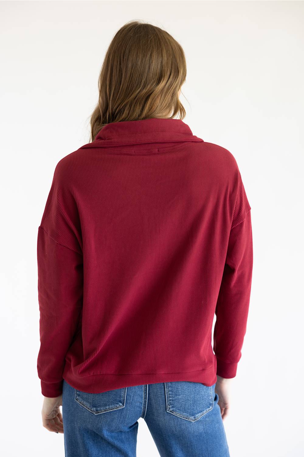 A woman viewed from behind wearing a dark red long-sleeve mock neck top and blue jeans against a plain white background.