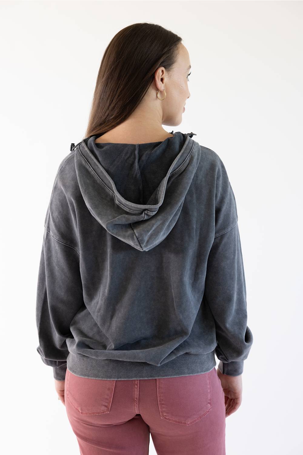 A woman shown from the back wearing a dark gray washed hooded sweatshirt and dusty rose pants against a plain white background.