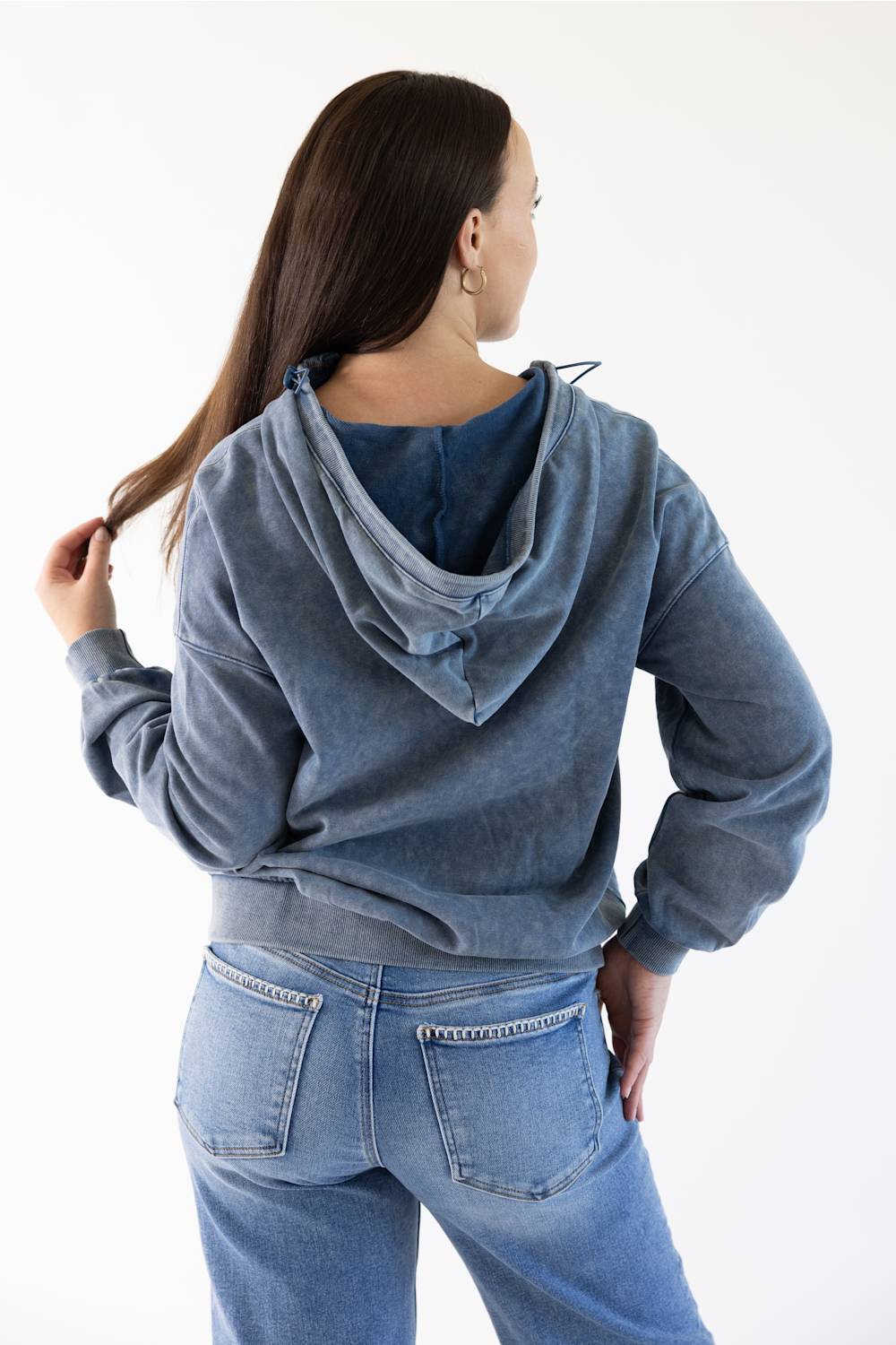 A woman shown from the back wearing a faded blue hooded sweatshirt and light blue jeans against a plain white studio background.
