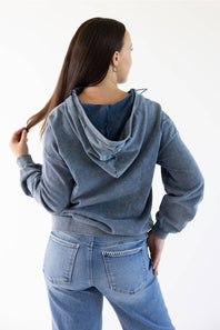 A woman shown from the back wearing a faded blue hooded sweatshirt and light blue jeans against a plain white studio background.
