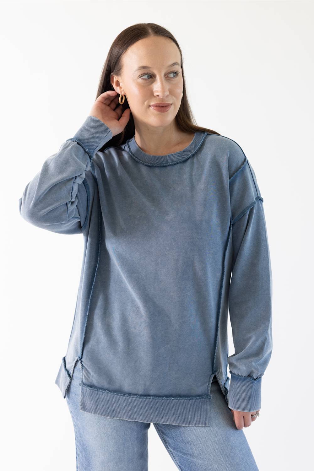 A woman modeling a washed slate blue long-sleeve top with unique gathered sleeve details and a relaxed silhouette, paired with blue jeans against a plain white studio background.