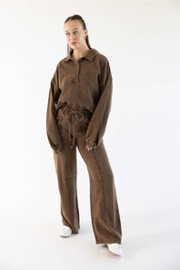 A woman modeling a matching brown two-piece lounge set consisting of a long-sleeve collared pullover and wide-leg drawstring pants, paired with white sneakers against a plain white studio background.