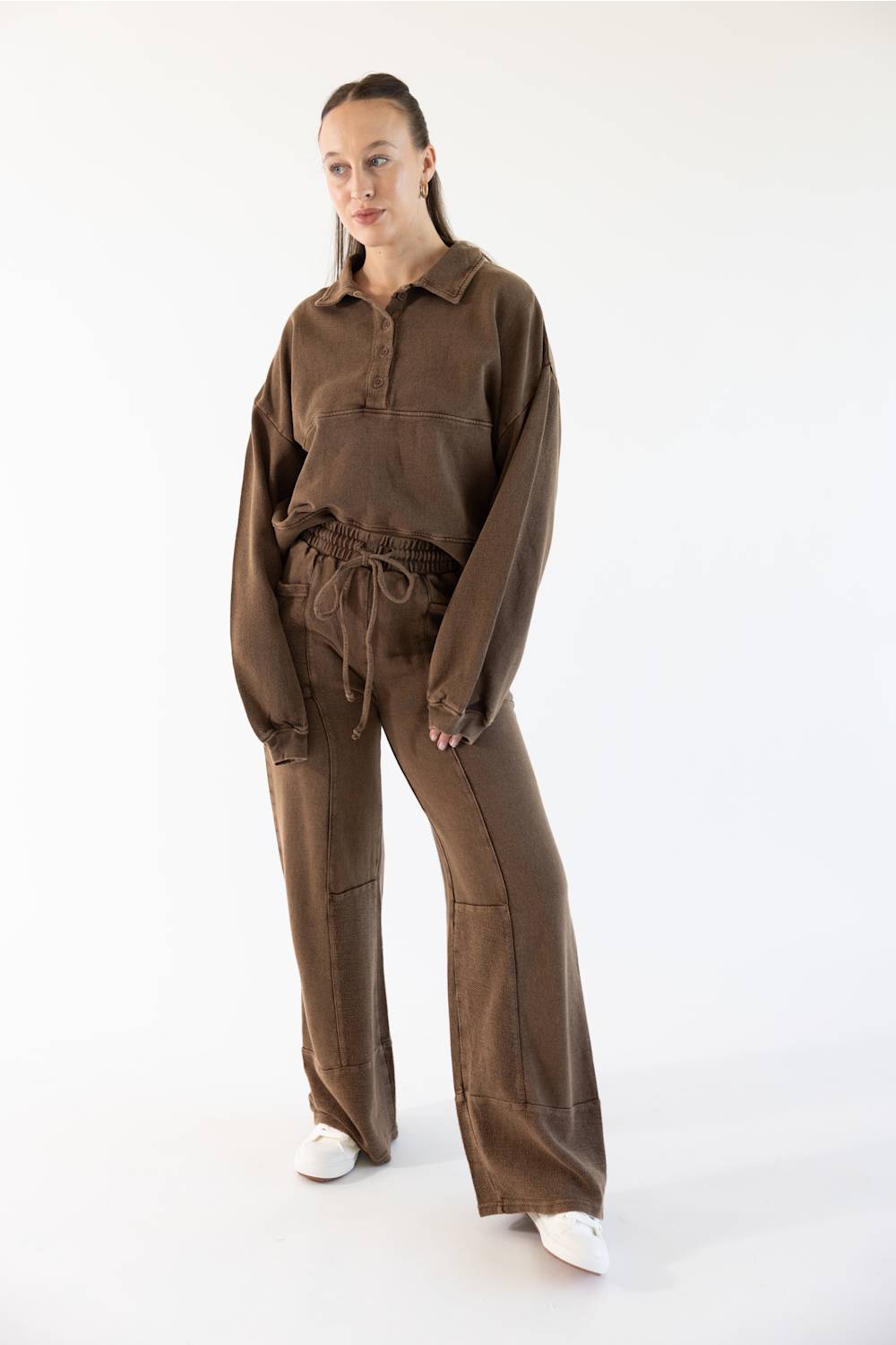 A woman modeling a matching brown two-piece lounge set consisting of a long-sleeve collared pullover and wide-leg drawstring pants, paired with white sneakers against a plain white studio background.