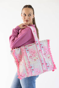 A woman wearing a purple sweatshirt and blue jeans carries a large woven tote bag with pink and white patterns and fringed edges against a plain white background.
