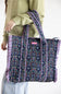A person wearing a sage green sweatshirt and light blue jeans carries a large pleated tote bag. The bag features a dark navy blue floral print with pink and purple striped ruffle trim along the edges and handles.