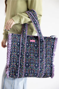 A person wearing a sage green sweatshirt and light blue jeans carries a large pleated tote bag. The bag features a dark navy blue floral print with pink and purple striped ruffle trim along the edges and handles.