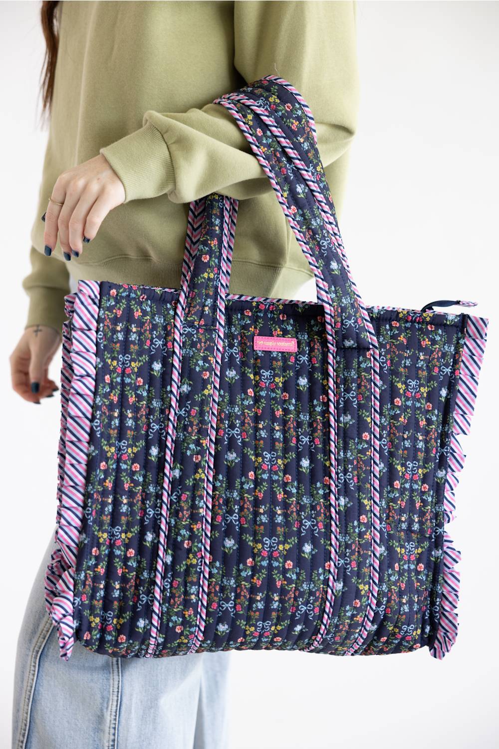 A person wearing a sage green sweatshirt and light blue jeans carries a large pleated tote bag. The bag features a dark navy blue floral print with pink and purple striped ruffle trim along the edges and handles.