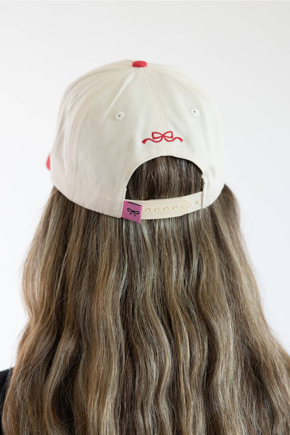 Back view of a model with long wavy hair wearing the Simply Southern Santa’s Favorite Holiday Hat for Women in Cream/Red, featuring a red embroidered bow above the keyhole cutout and an adjustable cream snapback closure with a pink bow tab.