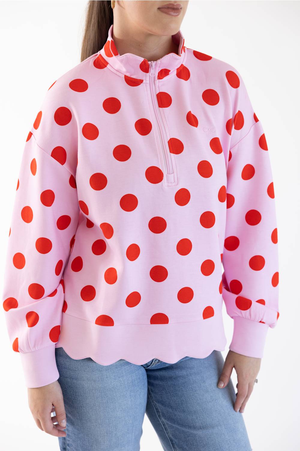 A woman wearing a pink long-sleeve quarter-zip pullover sweatshirt with large red polka dots and scalloped edge, paired with blue jeans, against a plain white background.