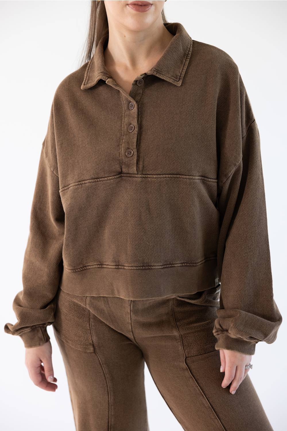 A woman wearing a brown cropped long-sleeve polo-style sweatshirt with a button placket and matching brown pants, shown against a plain light background.