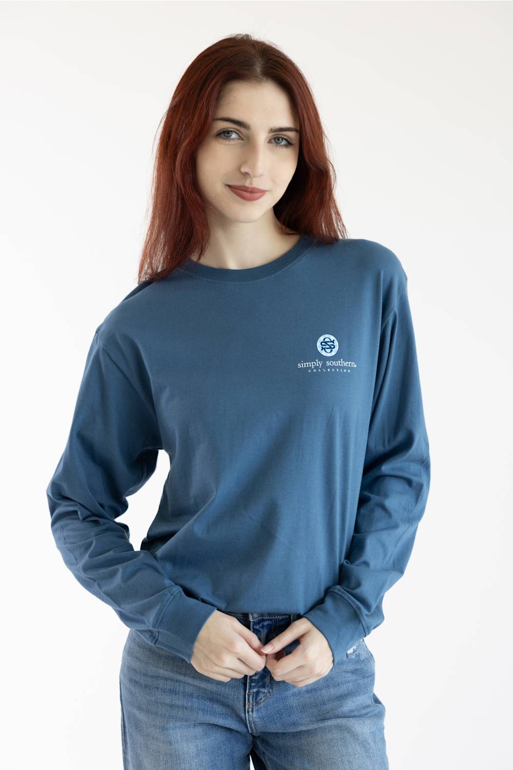 A woman with red hair wearing a slate blue long-sleeve crewneck sweatshirt with a small white circular logo on the left chest, paired with blue jeans, against a plain white studio background.