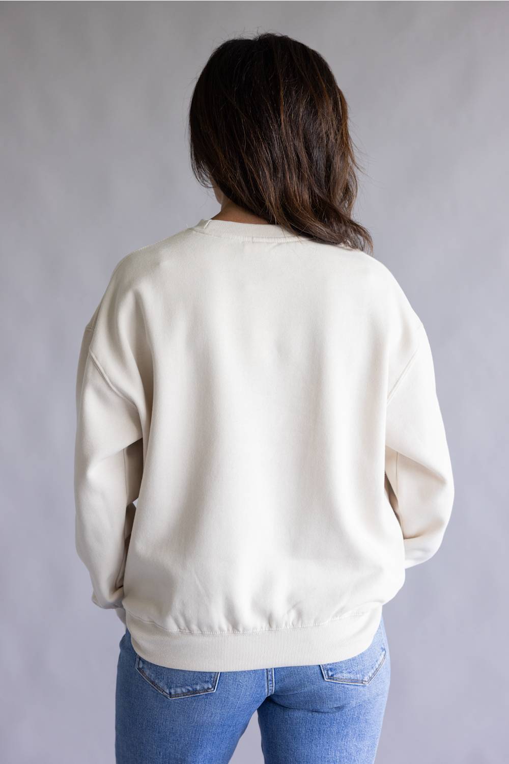 A person viewed from the back wearing an off-white crewneck sweatshirt and blue jeans against a plain gray studio background.