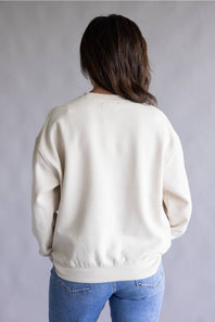 A person viewed from the back wearing an off-white crewneck sweatshirt and blue jeans against a plain gray studio background.