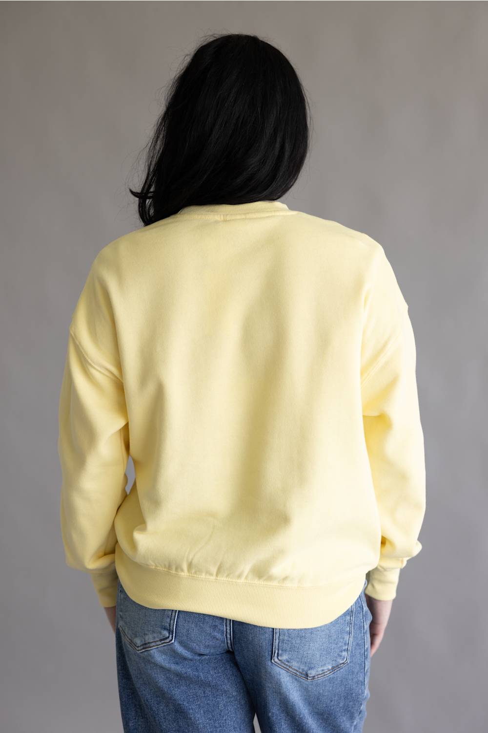 A person viewed from the back wearing a pale yellow crewneck sweatshirt and blue jeans against a plain gray studio background.