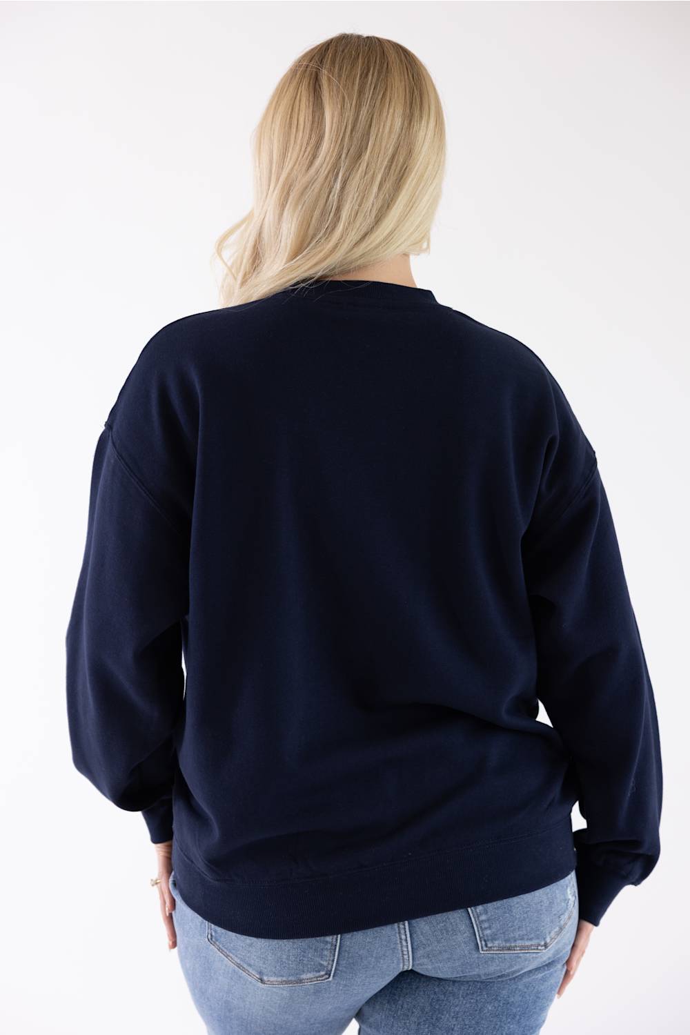 Back view of a blonde woman wearing a navy blue Reflex fleece crewneck sweatshirt and blue jeans.