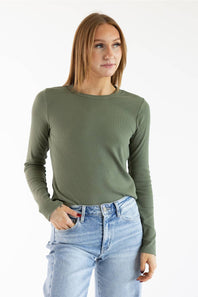 A woman wearing a fitted long-sleeve crewneck top in olive green, paired with light-wash blue jeans, photographed against a plain white studio background.