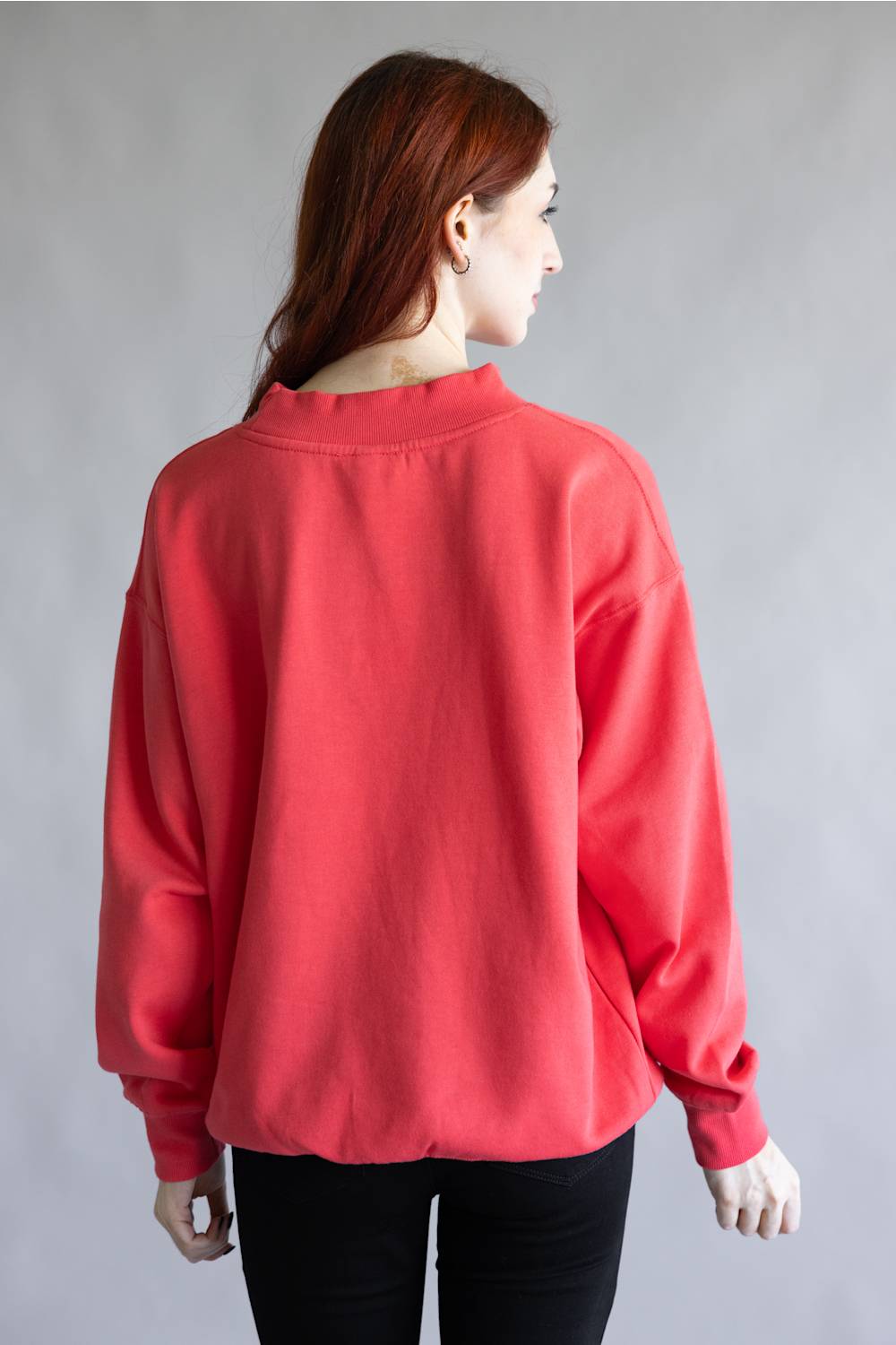 A woman with long auburn hair is shown from the back wearing a loose-fitting coral red sweatshirt and black pants against a plain gray studio background.