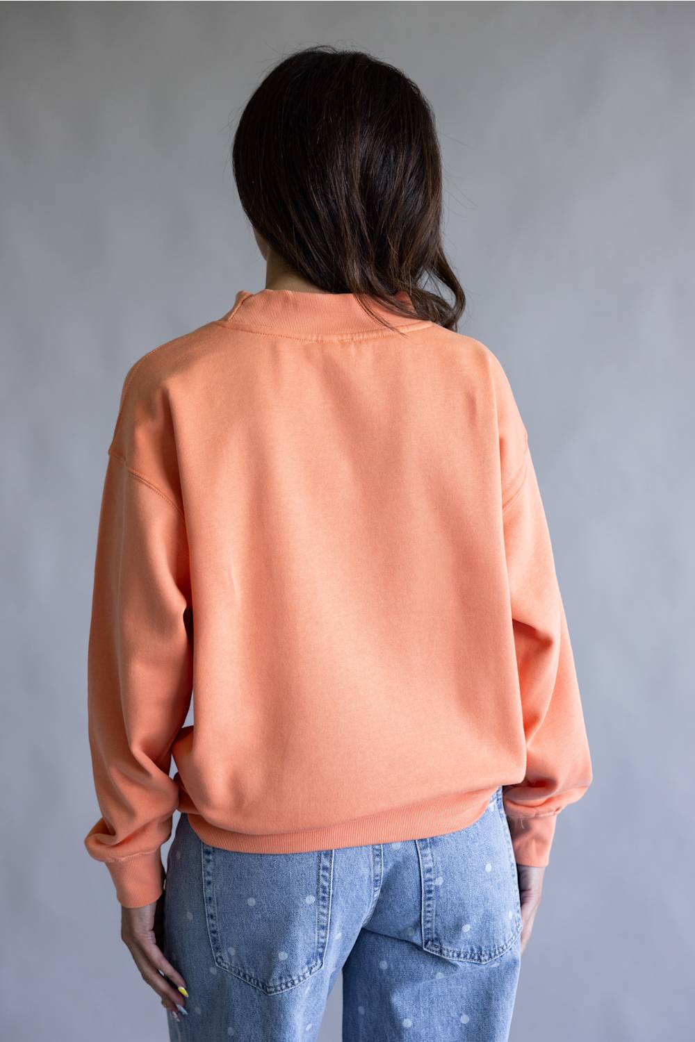 A person shown from the back wearing a peach-colored crewneck sweatshirt and light blue jeans, standing against a neutral gray background.