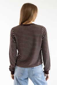 A woman viewed from behind wearing a brown and white striped long-sleeve shirt and light blue jeans against a plain white background.