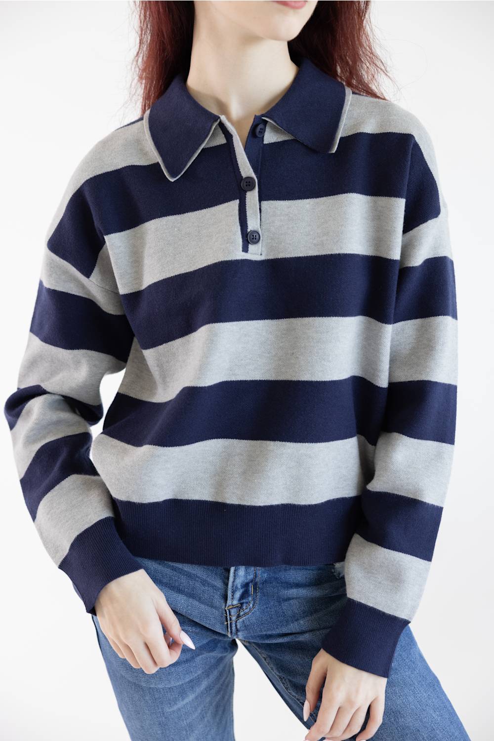A woman wearing a navy blue and light gray horizontally striped long-sleeve polo sweater with a three-button placket, paired with blue jeans, against a plain white background.