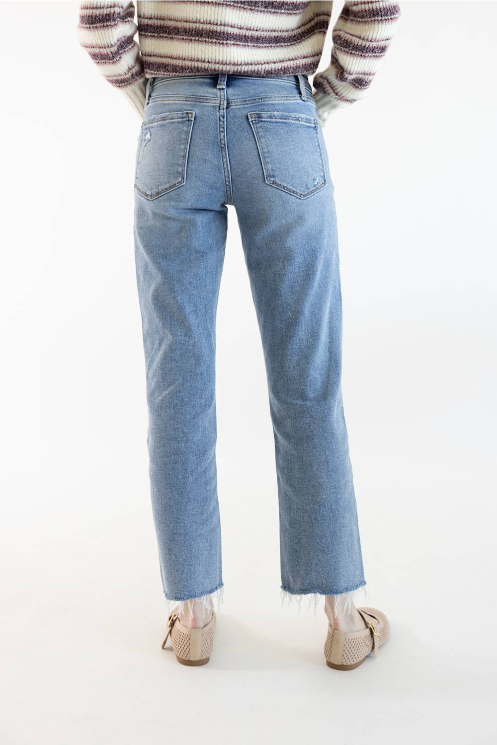 Rear view of a person wearing light-wash blue straight-leg jeans with a raw hem, paired with a striped sweater and beige loafers, against a plain white studio background.