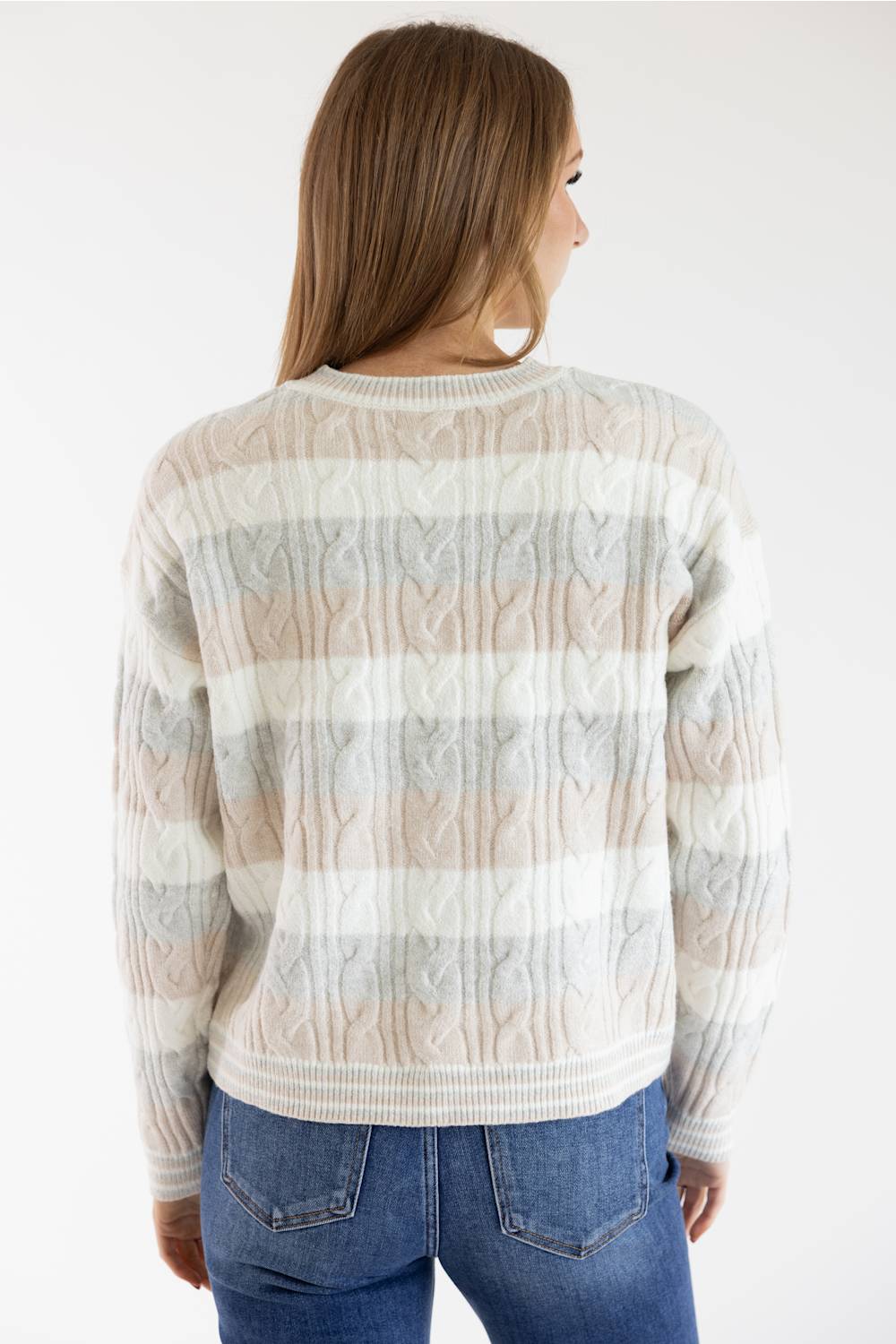 A woman viewed from the back wearing a cream, beige, and light gray cable-knit sweater with horizontal stripes, paired with blue jeans, against a plain white background.