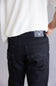 Close-up back view of a person wearing black denim jeans and a white textured top, showing the back pocket and a rectangular brand patch on the waistband.