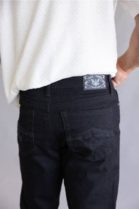 Close-up back view of a person wearing black denim jeans and a white textured top, showing the back pocket and a rectangular brand patch on the waistband.