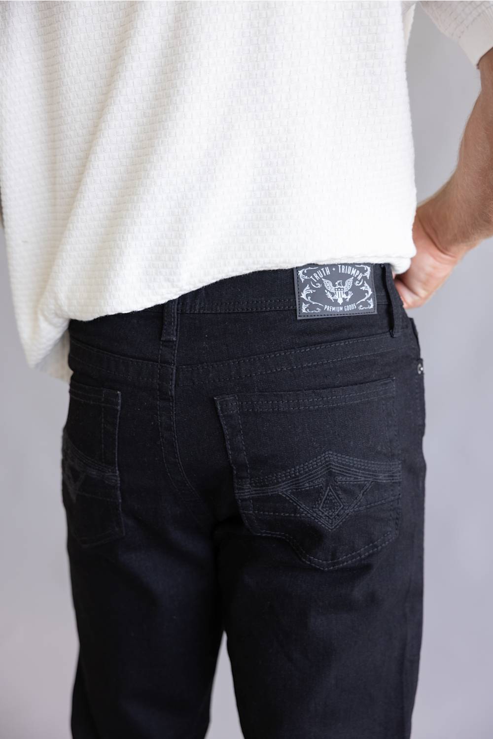 Close-up back view of a person wearing black denim jeans and a white textured top, showing the back pocket and a rectangular brand patch on the waistband.