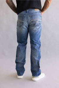 Rear view of a person wearing blue denim jeans with contrast stitching and white sneakers, standing against a light gray studio background.