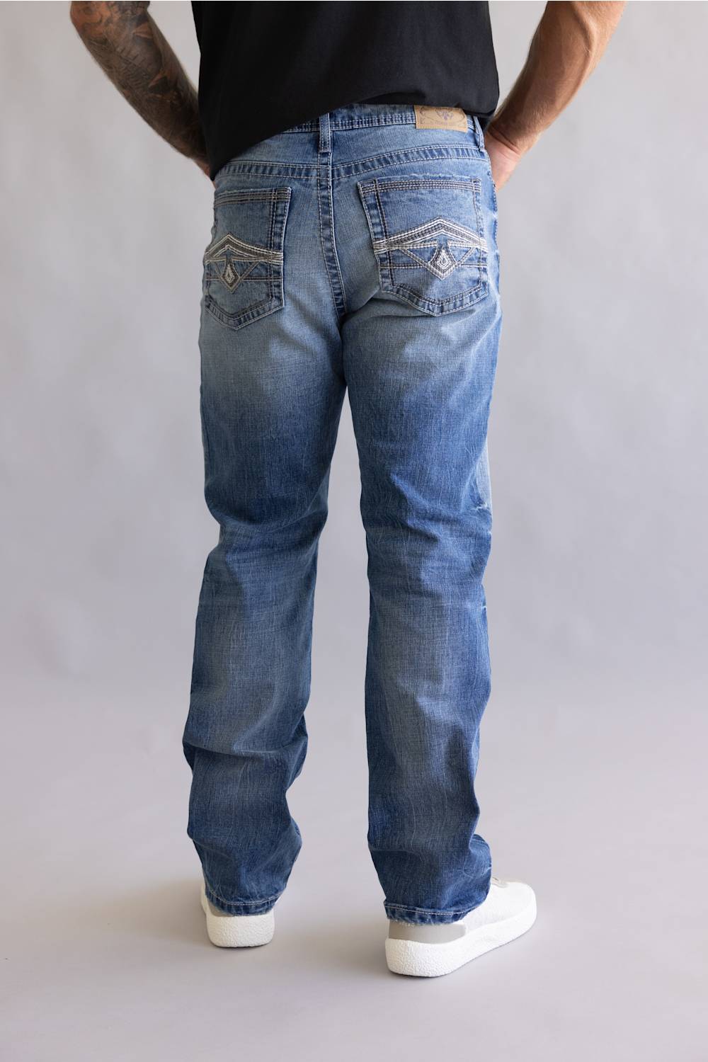 Rear view of a person wearing blue denim jeans with contrast stitching and white sneakers, standing against a light gray studio background.
