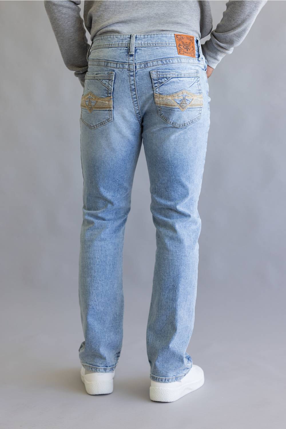 A person shown from the back wearing light-wash blue jeans with decorative gold embroidery on the back pockets, a gray long-sleeve top, and white sneakers against a neutral gray studio background.
