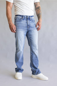 Rear view of a person wearing light blue denim jeans with decorative embroidered patch detailing on the back pockets, paired with a white t-shirt against a neutral gray background.