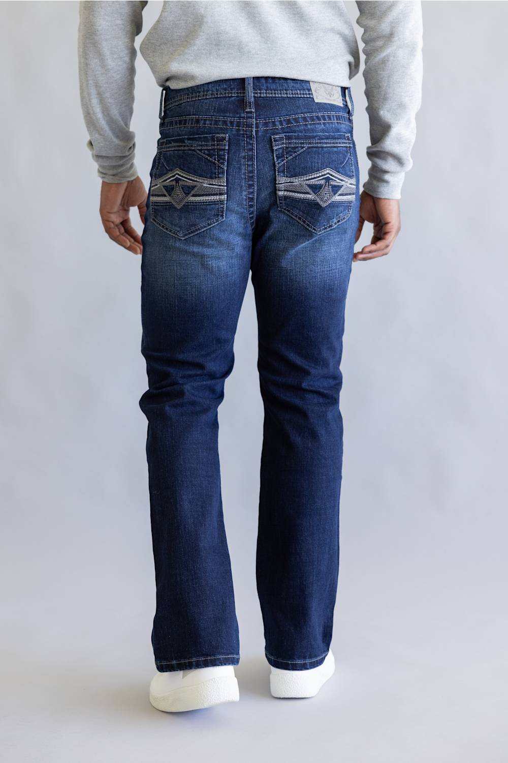 A person shown from the back wearing dark blue denim jeans with distinctive geometric embroidery on the back pockets, a light gray sweatshirt, and white sneakers, set against a plain light gray studio background.