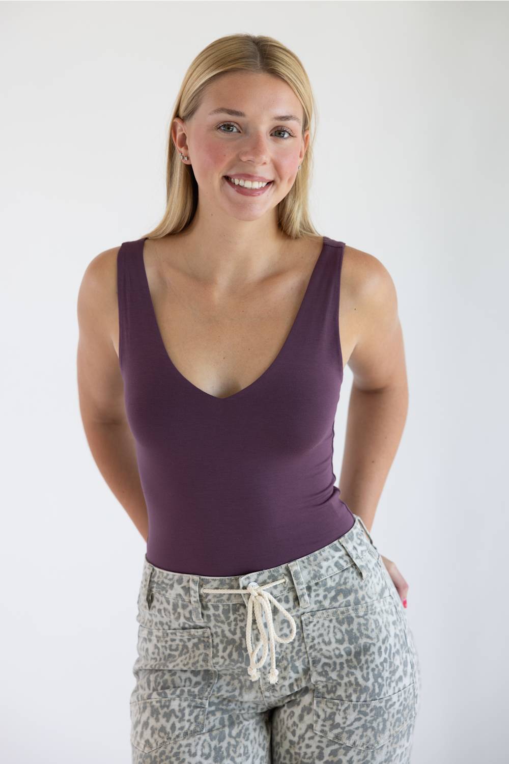 A smiling blonde woman wearing a sleeveless deep purple scoop-neck tank top and leopard-print pants, standing against a plain white studio background.