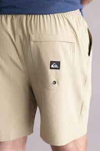 Close up of the back pocket of beige shorts with a brand logo on a plain background. The shorts are Quiksilver Taxer Amphibian 18" Hybrid Shorts for Men in Twill 