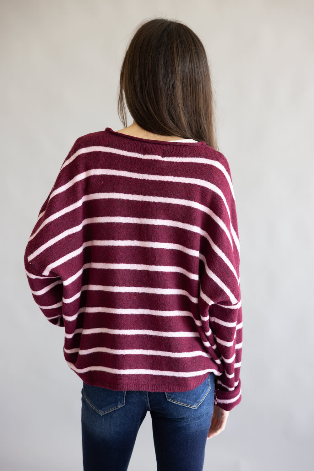 A woman viewed from behind wearing a loose-fitting, long-sleeve knit sweater with horizontal burgundy and light pink stripes, paired with blue jeans, against a plain light gray background.