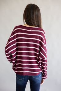 A woman viewed from behind wearing a loose-fitting, long-sleeve knit sweater with horizontal burgundy and light pink stripes, paired with blue jeans, against a plain light gray background.