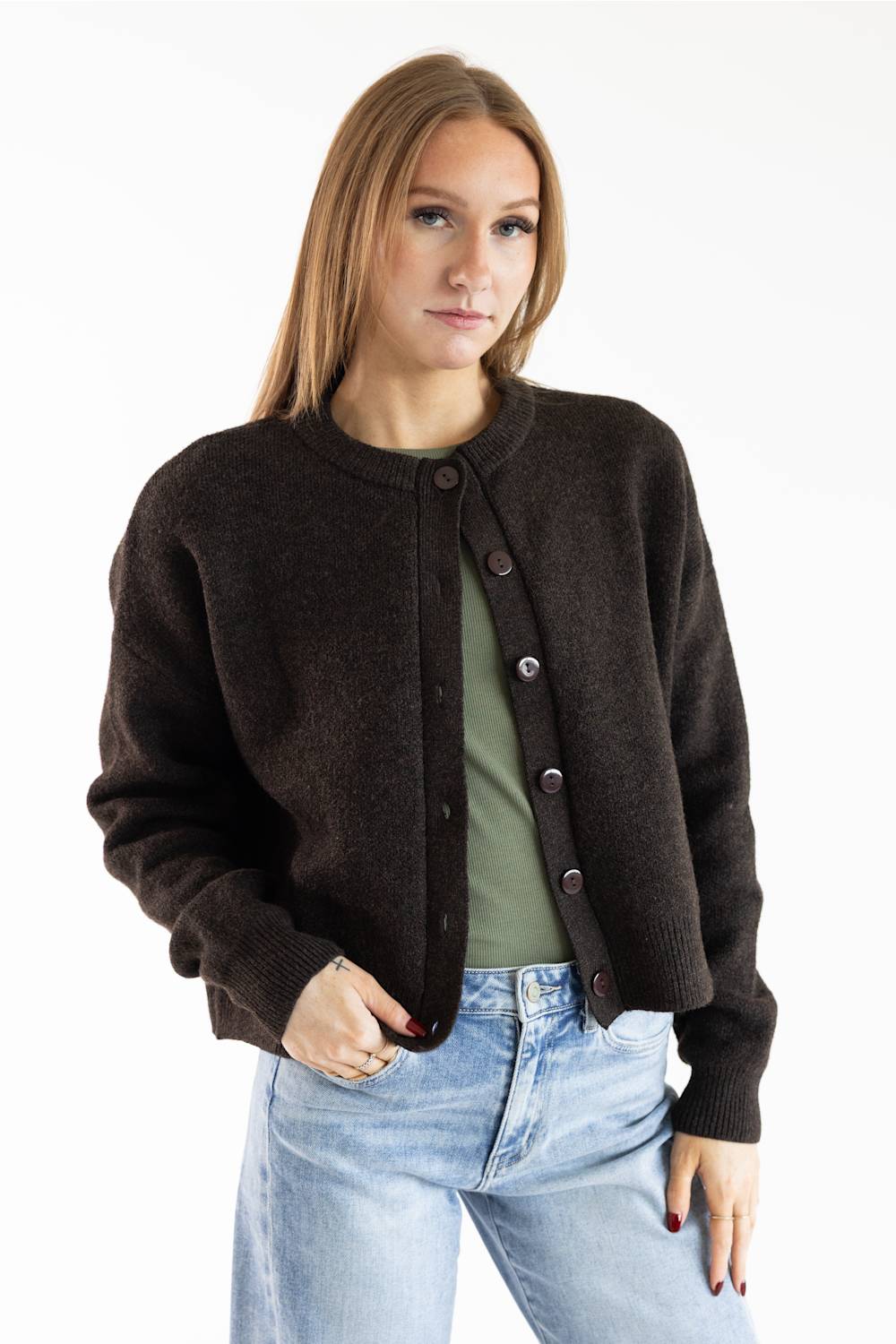 A woman modeling a dark brown knit cardigan with dark buttons over an olive green top and light blue jeans, shot against a plain white studio background.