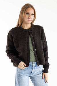 A woman modeling a dark brown knit cardigan with dark buttons over an olive green top and light blue jeans, shot against a plain white studio background.