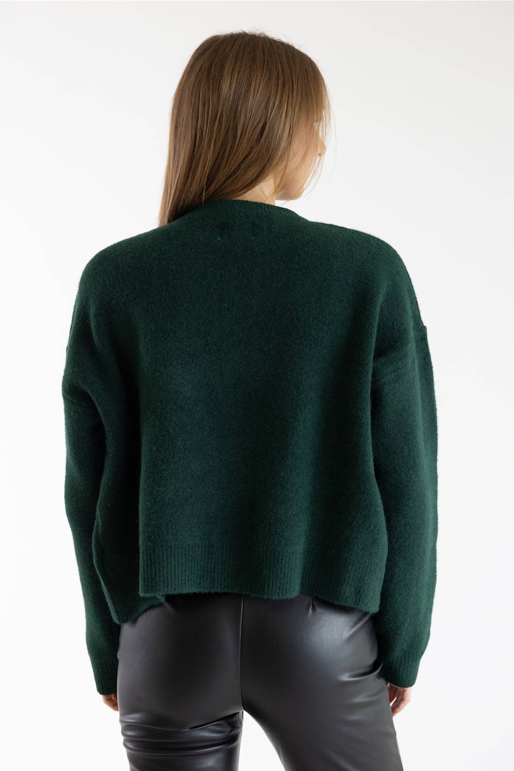 A woman viewed from the back wearing a dark forest green knit sweater and black leather pants against a plain white studio background.