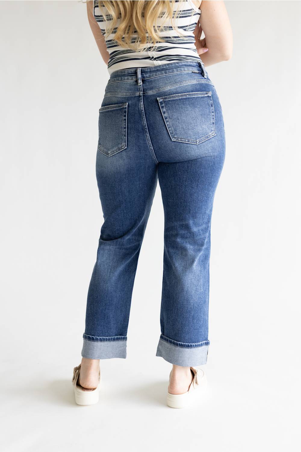 A person shown from the back wearing blue denim jeans with a cuffed hem, paired with a striped top and white platform sandals, against a plain light background.