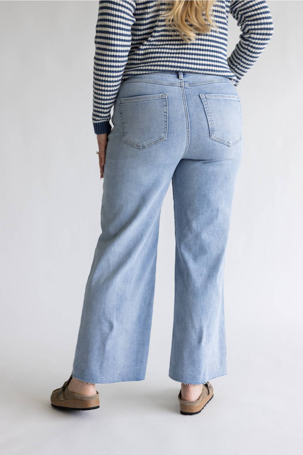 A person shown from the back wearing light-wash wide-leg denim jeans, a navy and white striped long-sleeve top, and tan flat sandals against a plain light gray studio background.