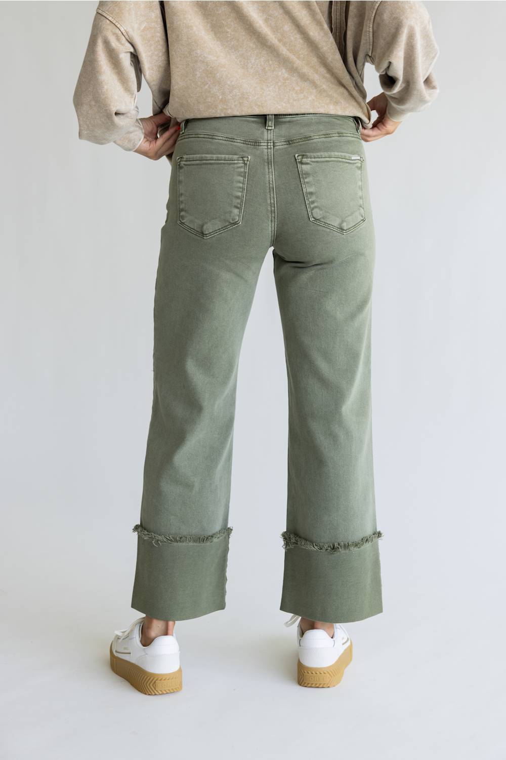 A person shown from the back wearing sage green wide-leg cropped jeans with rolled frayed hems, a beige sweater, and white platform sneakers against a light gray studio background.