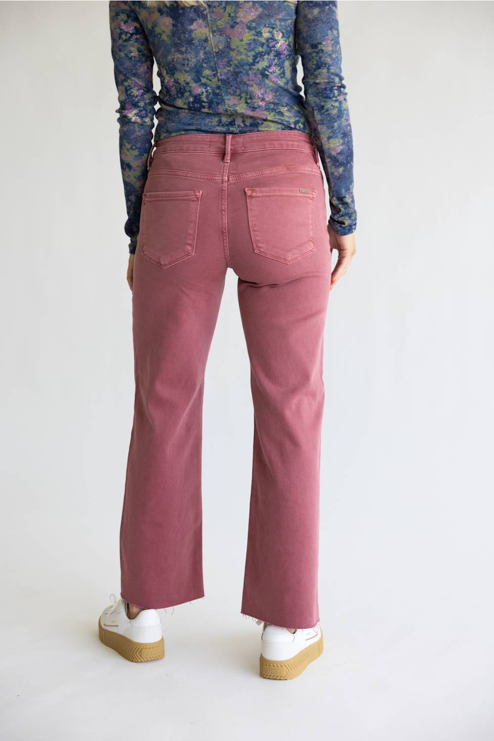 A person shown from the back wearing dusty rose flare jeans, a blue floral long-sleeve top, and white platform sneakers against a plain white studio background.