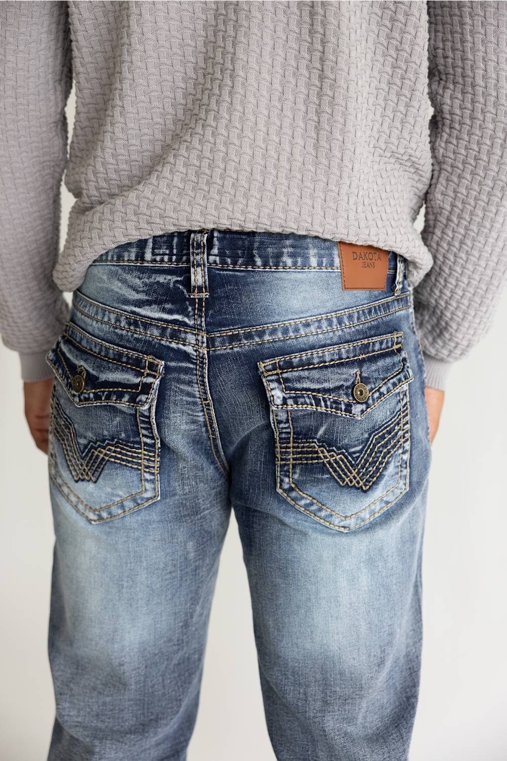Rear view of a person wearing light blue denim jeans with decorative contrast stitching and a brown leather patch on the back waistband, paired with a gray textured sweater.