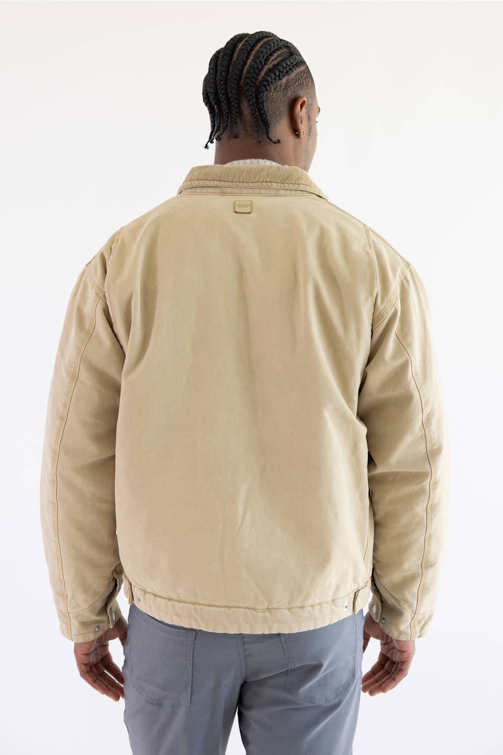 A man viewed from behind wearing a beige canvas utility jacket with a light-colored fleece collar and gray trousers, set against a plain white studio background.