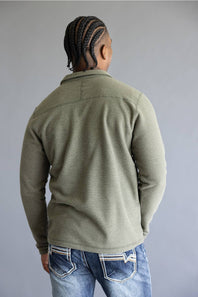 A man viewed from behind wearing an olive green long-sleeve knit sweater and blue jeans, standing against a plain gray studio background.