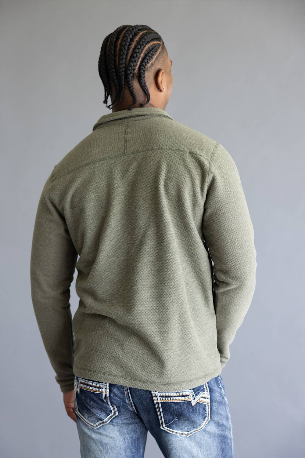 A man viewed from behind wearing an olive green long-sleeve knit sweater and blue jeans, standing against a plain gray studio background.