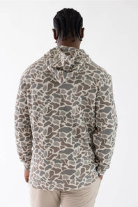 A man viewed from behind wearing a long-sleeve hooded sweatshirt with a camouflage pattern in beige, gray, and brown, paired with light beige pants against a plain white studio background.